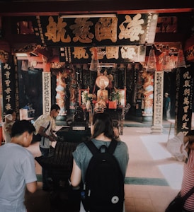 A traditional temple interior with ornate decorations, featuring a large statue at the back. Several people are interacting with objects in the foreground, one with a backpack. Incense coils hang from the ceiling, and there are intricate carvings and Chinese calligraphy on pillars and walls.