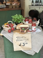 A rustic display of various food items including a basket of tomatoes, jars of tomato sauce, a loaf of bread, and a basil plant arranged on a table draped with cloth. A handwritten sign in the foreground advertises a promotion for £3.00.