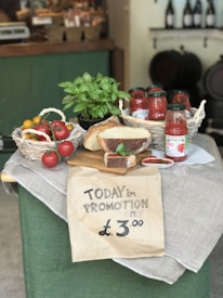A rustic display of various food items including a basket of tomatoes, jars of tomato sauce, a loaf of bread, and a basil plant arranged on a table draped with cloth. A handwritten sign in the foreground advertises a promotion for £3.00.