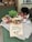 A rustic display of various food items including a basket of tomatoes, jars of tomato sauce, a loaf of bread, and a basil plant arranged on a table draped with cloth. A handwritten sign in the foreground advertises a promotion for £3.00.