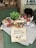 A rustic display of various food items including a basket of tomatoes, jars of tomato sauce, a loaf of bread, and a basil plant arranged on a table draped with cloth. A handwritten sign in the foreground advertises a promotion for £3.00.