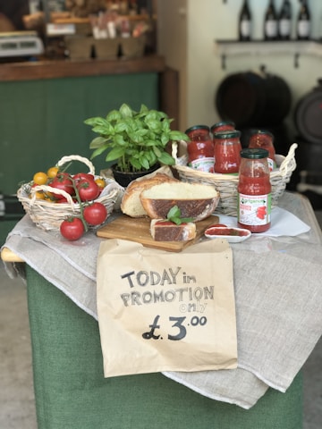 A rustic display of various food items including a basket of tomatoes, jars of tomato sauce, a loaf of bread, and a basil plant arranged on a table draped with cloth. A handwritten sign in the foreground advertises a promotion for £3.00.