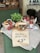 A rustic display of various food items including a basket of tomatoes, jars of tomato sauce, a loaf of bread, and a basil plant arranged on a table draped with cloth. A handwritten sign in the foreground advertises a promotion for £3.00.