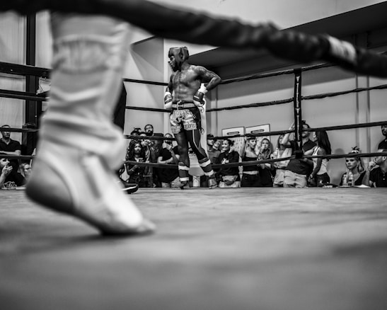 A boxer stands confidently in a ring, surrounded by an audience watching intently. The viewpoint is from behind the ropes, with a focus on the boxer wearing protective headgear and shorts, showing a sense of readiness and anticipation. The atmosphere is charged with energy, typical of a boxing event.