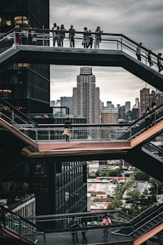 A modern architectural structure features multiple levels of walkways with people standing and walking. In the background, a city skyline with tall buildings is visible under a cloudy sky. The design includes geometric shapes and large glass windows.