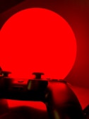A video game controller is silhouetted against a bright red circular background, creating a dramatic contrast. The image has a minimalistic composition with the controller featuring buttons and joysticks in shadow.