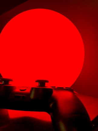 A video game controller is silhouetted against a bright red circular background, creating a dramatic contrast. The image has a minimalistic composition with the controller featuring buttons and joysticks in shadow.