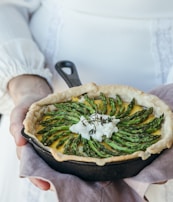 A rustic pie is garnished with neatly arranged asparagus spears and a dollop of cheese in a cast-iron skillet, resting on a cloth held by a person in a white, long-sleeved outfit.