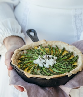 A rustic pie is garnished with neatly arranged asparagus spears and a dollop of cheese in a cast-iron skillet, resting on a cloth held by a person in a white, long-sleeved outfit.