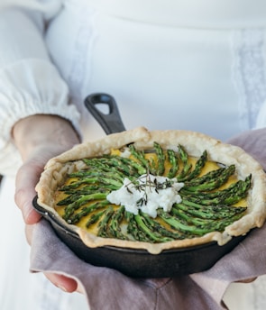 A rustic pie is garnished with neatly arranged asparagus spears and a dollop of cheese in a cast-iron skillet, resting on a cloth held by a person in a white, long-sleeved outfit.