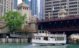A tour boat named 'Chicago's Little Lady' travels along a river, surrounded by the architectural features of a cityscape. There is a large bridge overhead with people observing the scene, and a lush green tree on the riverbank next to historic architectural structures.