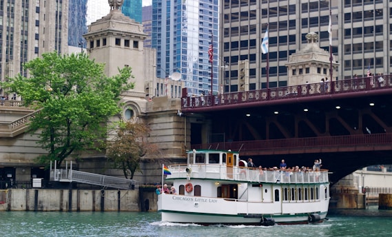 A tour boat named 'Chicago's Little Lady' travels along a river, surrounded by the architectural features of a cityscape. There is a large bridge overhead with people observing the scene, and a lush green tree on the riverbank next to historic architectural structures.