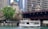 A tour boat named 'Chicago's Little Lady' travels along a river, surrounded by the architectural features of a cityscape. There is a large bridge overhead with people observing the scene, and a lush green tree on the riverbank next to historic architectural structures.