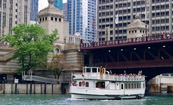 A tour boat named 'Chicago's Little Lady' travels along a river, surrounded by the architectural features of a cityscape. There is a large bridge overhead with people observing the scene, and a lush green tree on the riverbank next to historic architectural structures.
