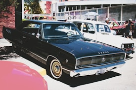 A classic black car parked in an outdoor setting, surrounded by other vintage vehicles. The scene includes people interacting with the cars, some taking photos. Buildings and trees are visible in the background, adding an urban feel to the environment.