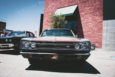 A classic car is prominently parked in front of a brick building on a sunny day. The vehicle has a vintage design with chrome details and distinctive round headlights. Another similar car is parked nearby, partially visible on the left.