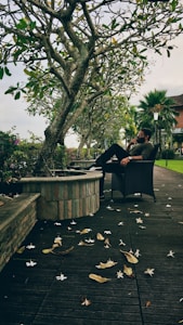 A man sits pensively on a wicker chair beside a stone planter under large leafy trees. Fallen leaves and white flowers are scattered across the wooden deck. The background shows a lawn with palm trees, a lamppost, and a building.