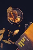 A cup of tea is placed on a saucer with a slice of ginger visible inside the cup. Dried leaves and cardamom are scattered around. There is a stack of books on the right side, with the top book featuring text in a non-Latin script and a vibrant yellow cover.