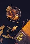 A cup of tea is placed on a saucer with a slice of ginger visible inside the cup. Dried leaves and cardamom are scattered around. There is a stack of books on the right side, with the top book featuring text in a non-Latin script and a vibrant yellow cover.