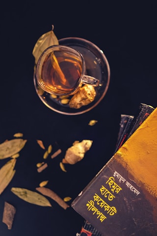 A cup of tea is placed on a saucer with a slice of ginger visible inside the cup. Dried leaves and cardamom are scattered around. There is a stack of books on the right side, with the top book featuring text in a non-Latin script and a vibrant yellow cover.