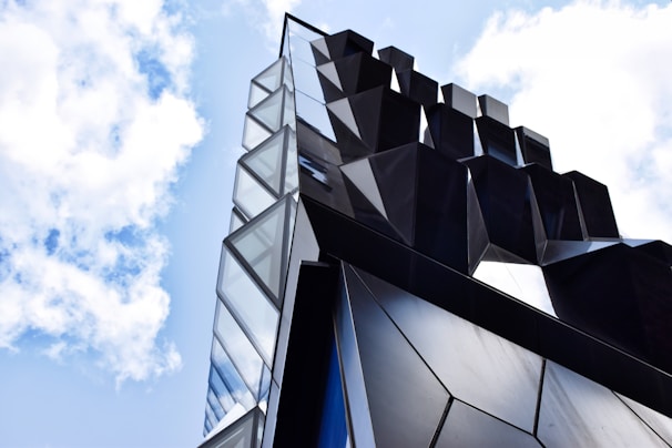 A modern architectural structure with a geometric design featuring large, angular glass and dark panels reaching into a bright blue sky with scattered clouds.