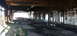 An abandoned industrial warehouse with a large open interior. The space is dimly lit, with beams of sunlight streaming in from the windows and holes in the roof. The floor is uneven and strewn with debris, puddles, and patches of moss. Rusted metal beams and supports run along the ceiling and walls, and graffiti is visible on the brick surfaces.