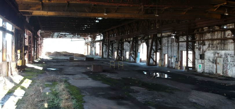 An abandoned industrial warehouse with a large open interior. The space is dimly lit, with beams of sunlight streaming in from the windows and holes in the roof. The floor is uneven and strewn with debris, puddles, and patches of moss. Rusted metal beams and supports run along the ceiling and walls, and graffiti is visible on the brick surfaces.