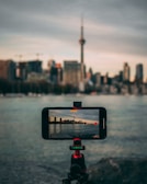 A smartphone mounted on a tripod is centered in the foreground, capturing a photo of a city's skyline across a body of water. A prominent tower can be seen among the buildings, and the sky is overcast.