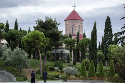 A church with a red-tiled roof is surrounded by lush green foliage and various tall trees. Two people in dark clothing are walking away on a pathway beside manicured lawns and steps. The sky is overcast, creating a serene and peaceful atmosphere.