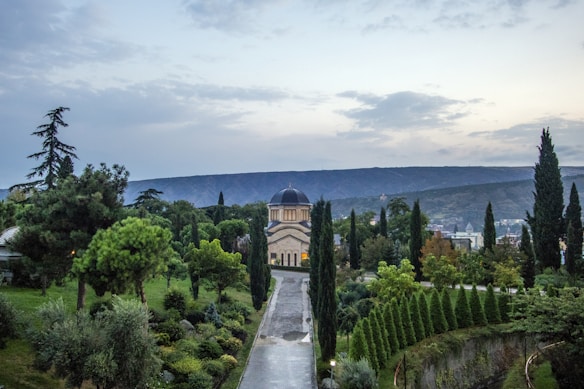A peaceful landscape features a pathway lined with trees leading to a domed building in the distance. Surrounding the path are lush, well-maintained gardens filled with a variety of trees and shrubs. The landscape is framed by hills under a partly cloudy sky at dusk.
