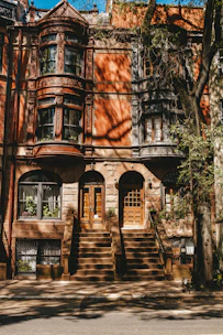 A classic brownstone building with intricate architectural details and large bay windows. The building has two sets of stone steps leading to wooden double doors. There are shadows of trees cast on the facade, and some greenery is visible next to the steps and in the windows.