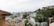 A hillside neighborhood overlooking a city skyline. The scene includes a variety of houses in different architectural styles, interspersed with lush greenery and palm trees. In the background, modern high-rise buildings are visible, and the sky is slightly overcast with scattered clouds.