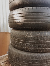 Four stacked, worn-out car tires are placed on a wooden pallet with a corrugated metal wall in the background.
