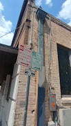 An exterior corner of a brick building with several signs attached to a metal post. One sign indicates 'No Parking Musician Loading 7 PM - 3 AM', and another sign indicates '30 Min Commercial Zone 9 AM - 7 PM'. There is also a sign showing a tow-away zone. Electrical boxes and conduits are mounted on the wall.
