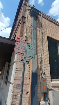 An exterior corner of a brick building with several signs attached to a metal post. One sign indicates 'No Parking Musician Loading 7 PM - 3 AM', and another sign indicates '30 Min Commercial Zone 9 AM - 7 PM'. There is also a sign showing a tow-away zone. Electrical boxes and conduits are mounted on the wall.