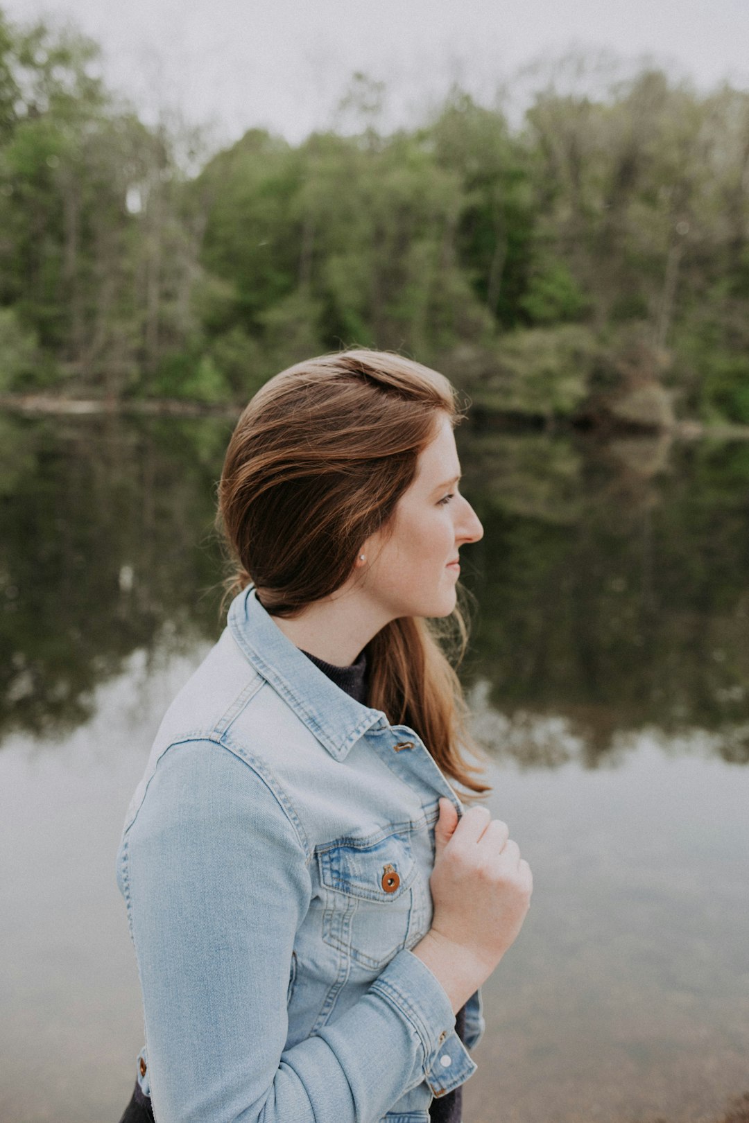 woman in faded blue denim jacket standing in bank of lake