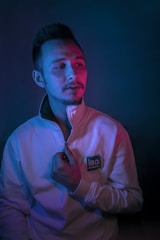 A person wearing a zip-up sweater stands against a dark backdrop, illuminated by contrasting blue and pink lighting. The sweater has a logo that reads 'IBC Engineering.' The individual appears contemplative, holding the zipper with one hand.