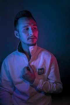 A person wearing a zip-up sweater stands against a dark backdrop, illuminated by contrasting blue and pink lighting. The sweater has a logo that reads 'IBC Engineering.' The individual appears contemplative, holding the zipper with one hand.
