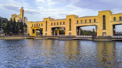 A large, historic building with an ornate facade sits alongside a body of water. The structure appears to be a hydroelectric dam, characterized by its distinctive architectural design, including arches and a mix of modern and classical elements. In the background, another building with a tower is visible, surrounded by greenery.