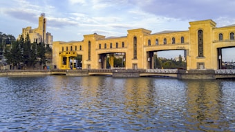 A large, historic building with an ornate facade sits alongside a body of water. The structure appears to be a hydroelectric dam, characterized by its distinctive architectural design, including arches and a mix of modern and classical elements. In the background, another building with a tower is visible, surrounded by greenery.