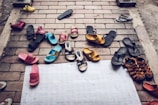 A variety of sandals and flip-flops are scattered on a brick pavement, including different colors like black, blue, pink, yellow, and brown. Some of the sandals have playful designs, while others are more plain. The arrangement appears random, suggesting a cluttered or casual scene.