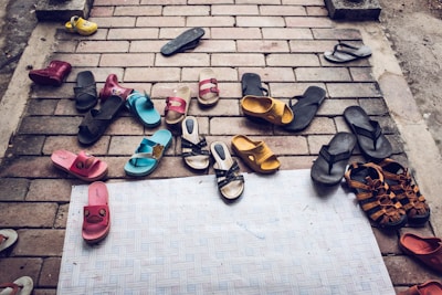 A variety of sandals and flip-flops are scattered on a brick pavement, including different colors like black, blue, pink, yellow, and brown. Some of the sandals have playful designs, while others are more plain. The arrangement appears random, suggesting a cluttered or casual scene.