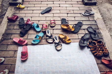 A variety of sandals and flip-flops are scattered on a brick pavement, including different colors like black, blue, pink, yellow, and brown. Some of the sandals have playful designs, while others are more plain. The arrangement appears random, suggesting a cluttered or casual scene.