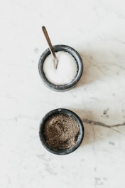 Two small stone bowls are placed on a light marble surface. The top bowl contains a coarse white substance and has a metal spoon in it, likely containing salt. The bottom bowl is filled with a dark, granular substance, likely pepper.