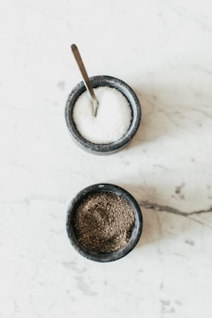 Two small stone bowls are placed on a light marble surface. The top bowl contains a coarse white substance and has a metal spoon in it, likely containing salt. The bottom bowl is filled with a dark, granular substance, likely pepper.