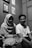 A black and white photo featuring two people sitting closely together. The person on the left is wearing a headscarf and a striped shirt, and the person on the right is in a plain t-shirt. They are both smiling. The room has wooden walls with framed photos hanging.