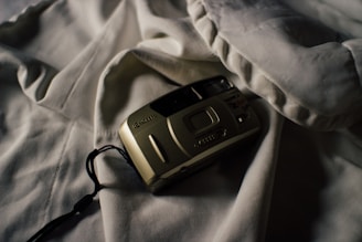 A vintage film camera rests on a soft, rumpled white fabric, casting a shadow. The lighting creates a moody and nostalgic atmosphere, highlighting the contours of the camera.