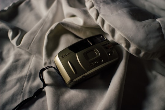 A vintage film camera rests on a soft, rumpled white fabric, casting a shadow. The lighting creates a moody and nostalgic atmosphere, highlighting the contours of the camera.
