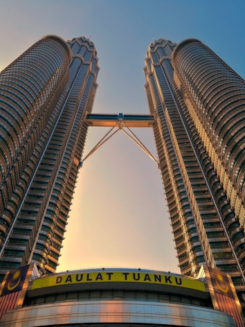 Two tall, identical skyscrapers connected by a sky bridge dominate the image. The buildings feature a modern architectural design with numerous glass windows and metallic elements. At the base, a banner displays the words 'DAULAT TUANKU' in bold yellow letters, accompanied by the Malaysian flag with its distinctive crescent and star.