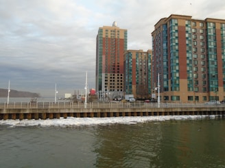 High-rise buildings with blue and red accents stand adjacent to a body of water. The waterfront area has a promenade with railings, and the water appears calm with a few visible boats and sparse traffic on the road.